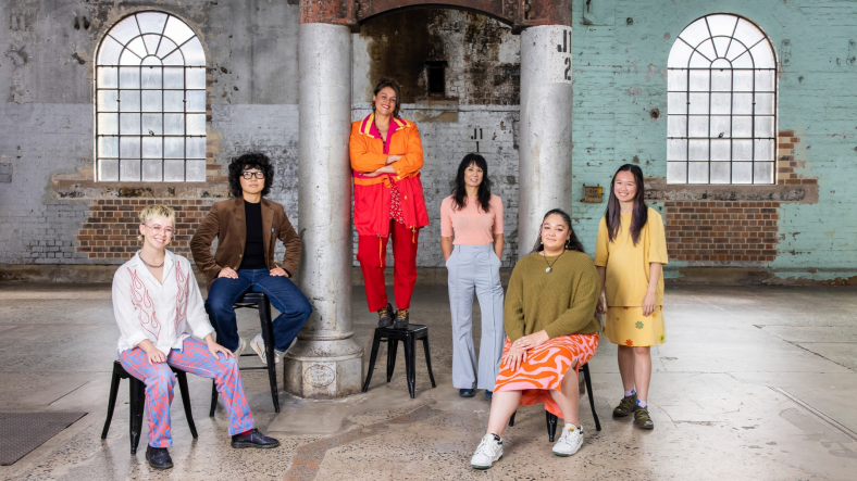 A group of young, colourfully dressed artists pose against interior Carriageworks backdrop.