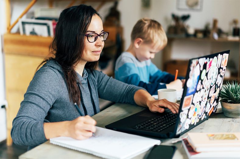 A close-up shot of a mother wearing a grey shirt and black glasses working on a laptop with her toddler son sitting beside her