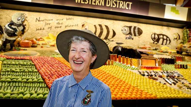 smiling older woman wearing a hat standing in front of a display of fresh fruit
