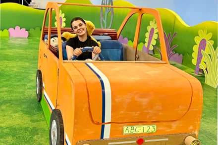 Young woman smiling, seated in big orange cardboard car on colourful Playschool set