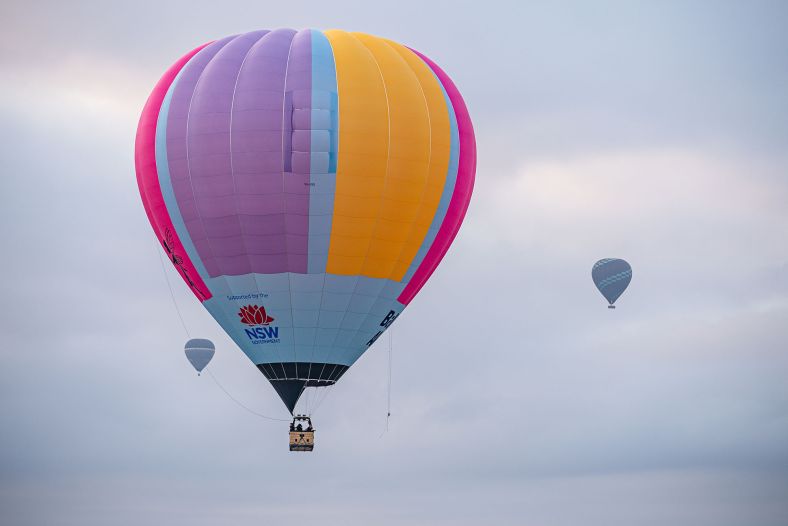 Three hot air balloons float in the sky. The one in the foreground is purple, pink, blue and orange and has supposrted by the NSW Government written on the side. The two in the background are grey. 