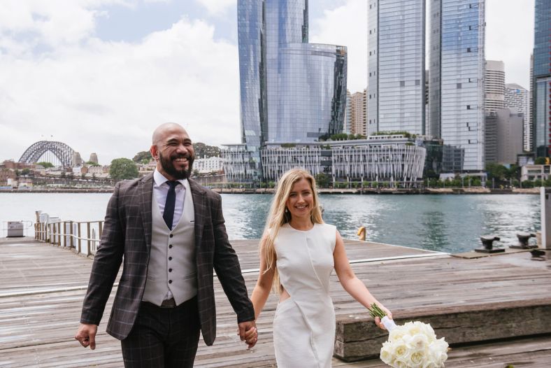 A bride and groom walk along a harbourside wharf. Behind them you can see the city skyline and Sydney Harbour Bridge.