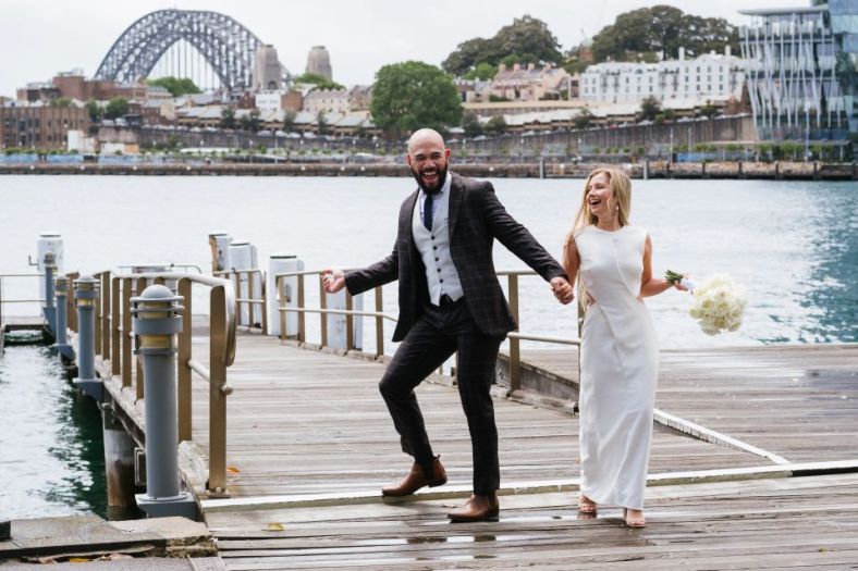 A bride and groom walk as if they are dancing on a harbourside wharf. In the background is the Sydney Harbour Bridge.