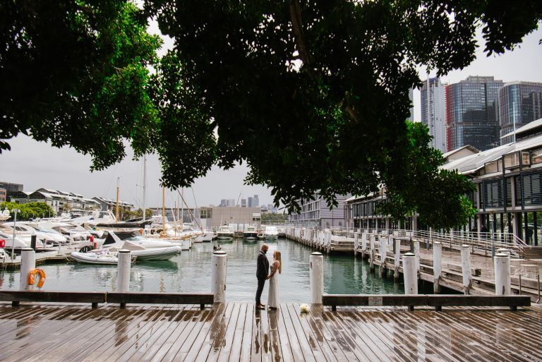 A bridge and groom look pose for the camera framed by deep green tree canopy. Behind them boats are moored on the wharf
