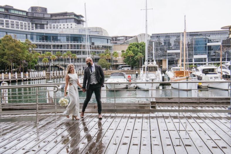 A bride and groom walk along a wooden wharf shiny with recent rain. Behind them boats are moored and the Pyrmont harbourside buildings reflect the light off the water.