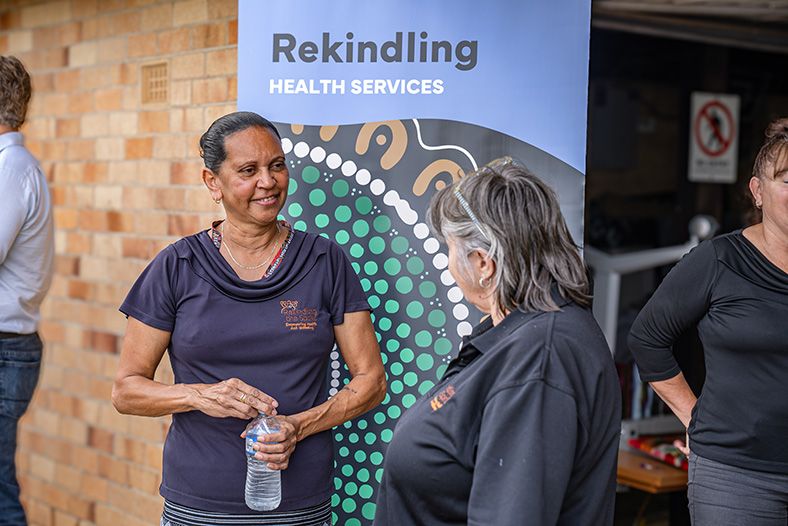 Two women talking in front of a "Rekindling health services" banner, wearing "Rekindle the Spirit" t-shirts in Lismore at the announcement of funding through the Aboriginal-owned Assets Program.