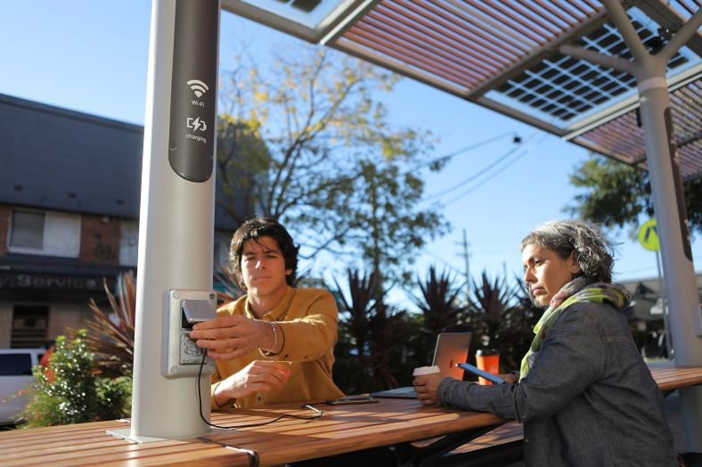 Two people are sitting outside at a ChillOUT Hub in the Georges River Council area, using the provided wi-fi and powerpoints. Credit: Georges River Council