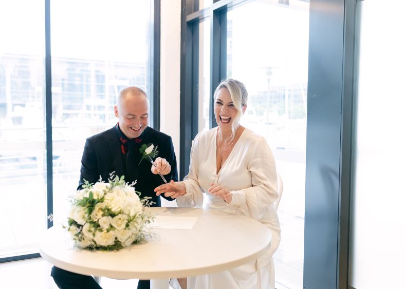 A couple sits a table by a bright window to sign their marriage certificate. They look very happy and one is laughing.