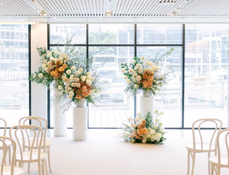 a brightly lit room with chairs forming an aisle for a wedding ceremony. Flowers are set up at the end of the aisle