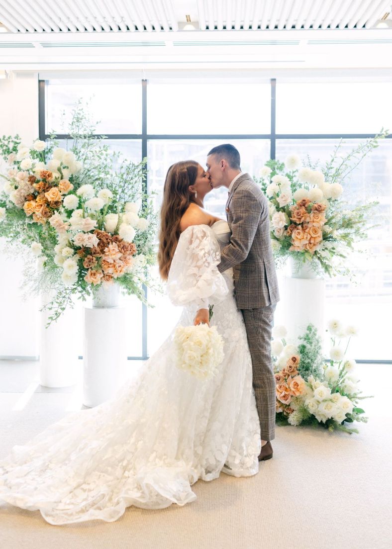 A bride and groom in a brightly lit room share a kiss at the end of the aisle. They are framed with flowers in the background.