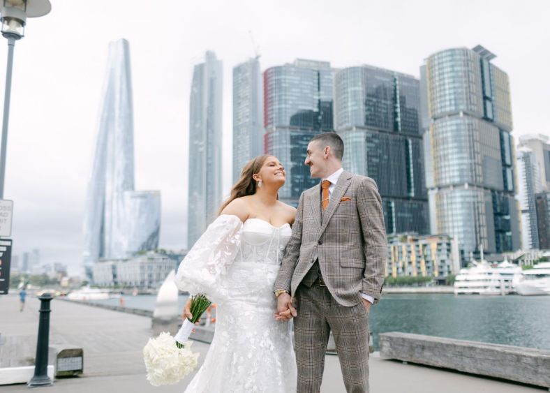 A bride and groom smile at each other on a city wharf. Behind them the city is bright silver.