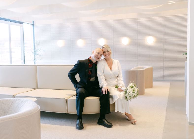 A couple poses for the camera while they sit on modern lounges inside the Registry. The colours around them are creams and the room is filled with light.