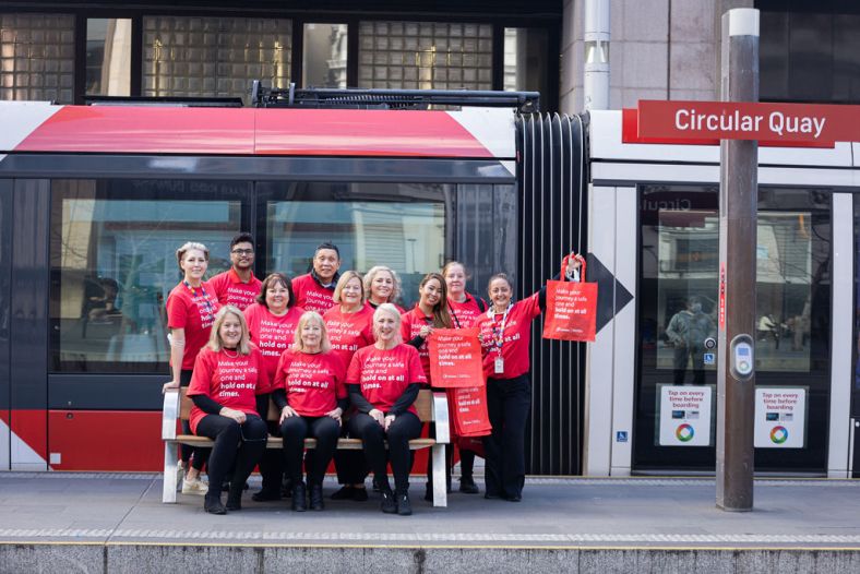 Group of people sitting in front of circular quay light rail smiling to camera in red