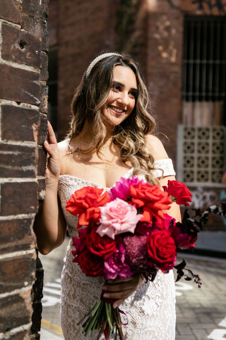 Smiling bride standing next to a brick wall holding a bouquet of red and pink flowers.