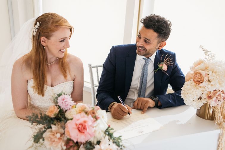 Bride and groom sitting at a table looking at each other while groom signs a certificate.