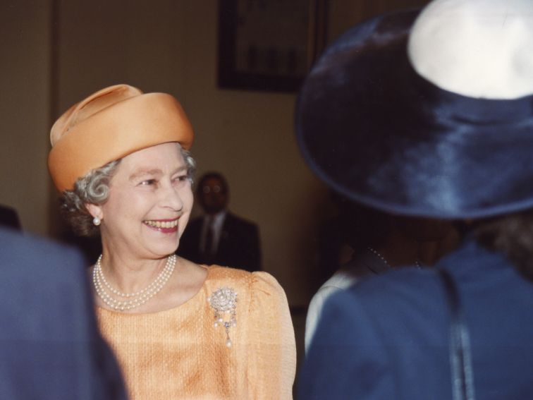 The Queen in an apricot coloured hat and jacket, smiling at someone out of shot. 