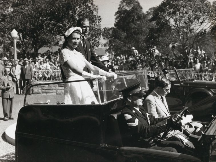  Black and white photo of the Queen and Duke of Edinburgh riding through the streets of Sydney standing on the back of a car. 