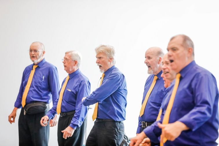 Group of senior performers in blue shirts and yellow ties singing with enthusiasm at Seniors Festival