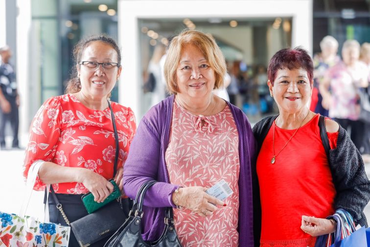 Three women huddle close and smile for camera outside Seniors Expo or Festival event