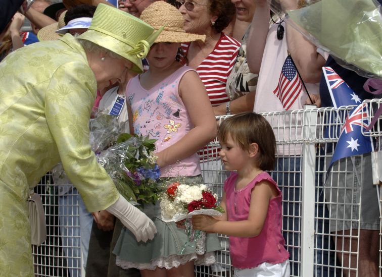 Her Majesty Queen Elizabeth II is bending down to receive flowers from a toddler. Her Majesty is wearing a light lime green skirt and jacket with a matching hat. 