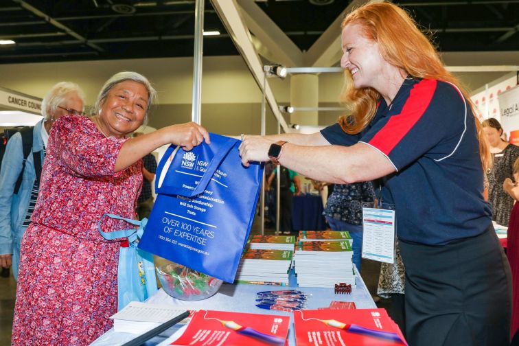 Young woman hands Seniors Expo attendee a show bag over counter