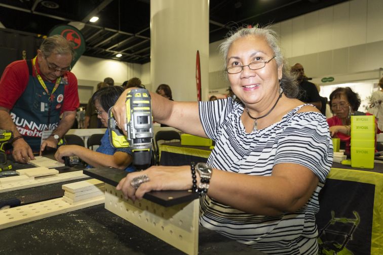 Woman holding hand drill above wood and smiling at Bunnings workshop Seniors Festival Expo