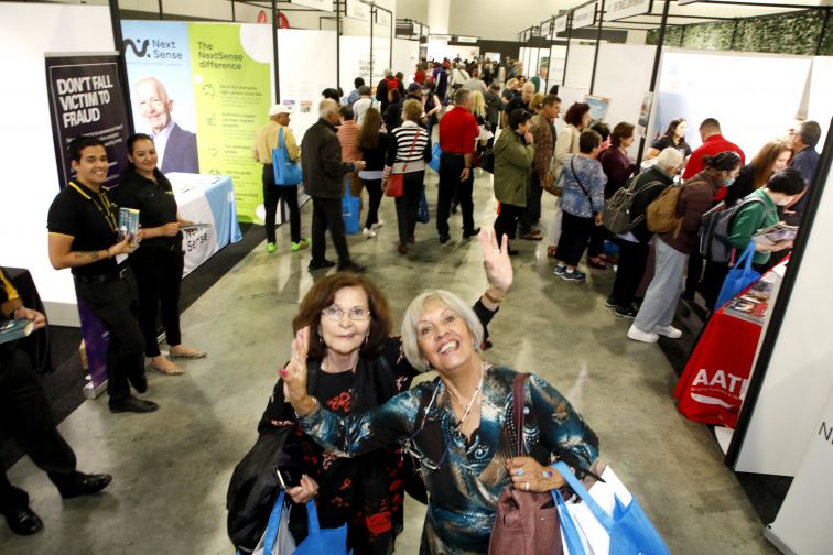 Ladies at Seniors Expo happily waving at camera, while clutching 'showbags', with crowd milling in background
