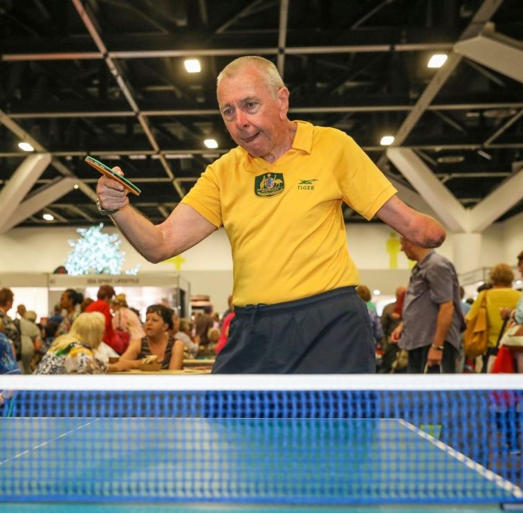 Older man with raised table tennis bat as he returns ball over net with focus aat Seniors Festival Expo