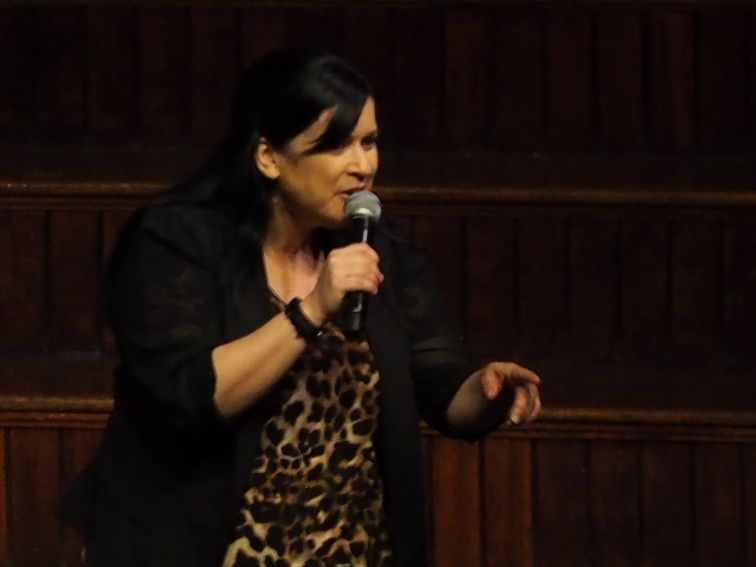 Female comedian dressed in leopard spot shirt and black jacket telling audience a funny story