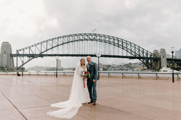 A couple embrace on the Sydney Opera House forecourt with the harbour bridge in the background