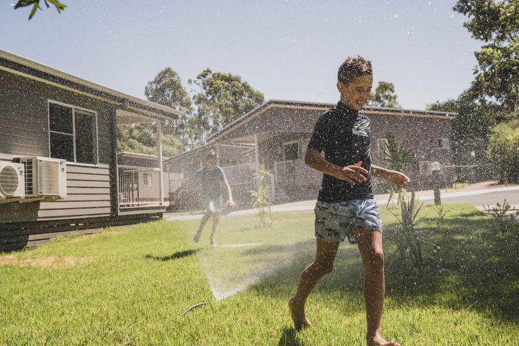 Children run through a water sprinkler outdoors on a lawn, next to a house.