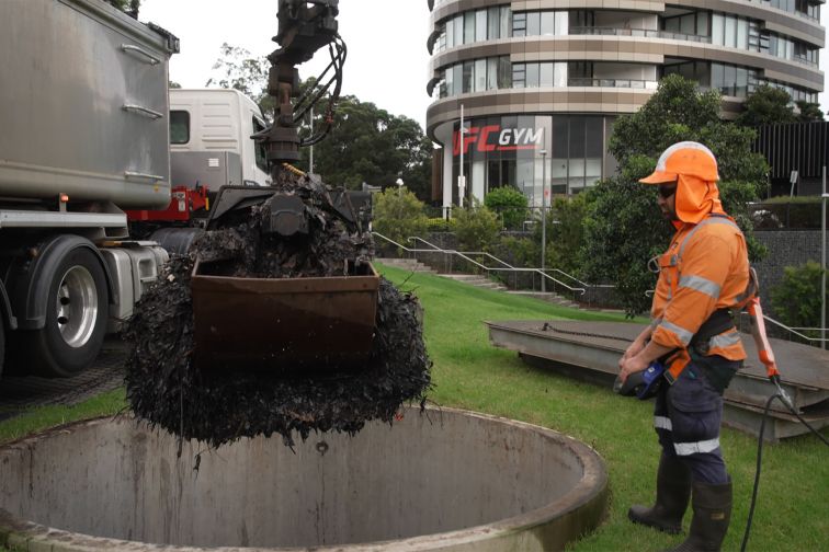 A man in high visibility clothing and helmet operates a crane, cleaning out a gross pollutant trap in Sydney Olympic Park.