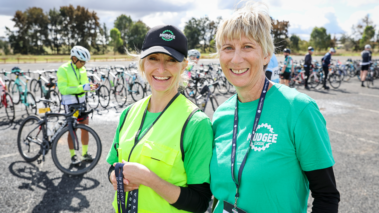 two smiling volunteers with cyclists behind them at the mudgee classic