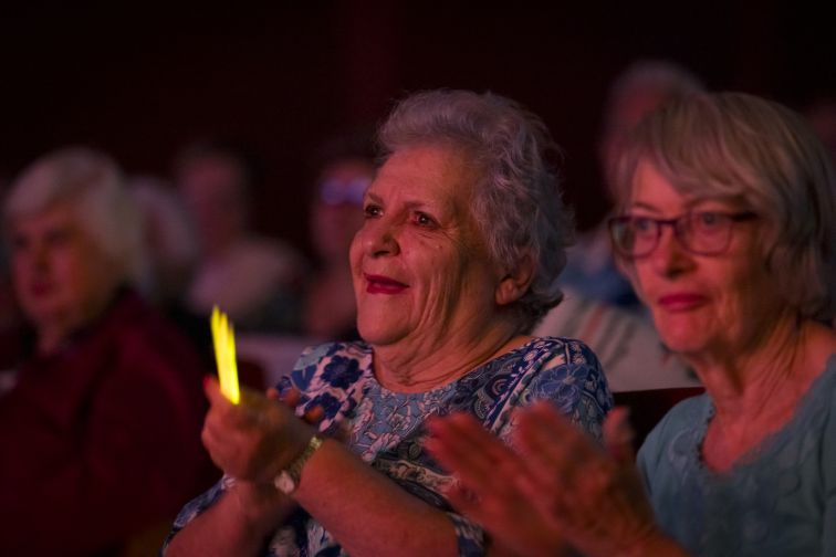 Older woman sitting in audience, holding a glowstick and smiling.