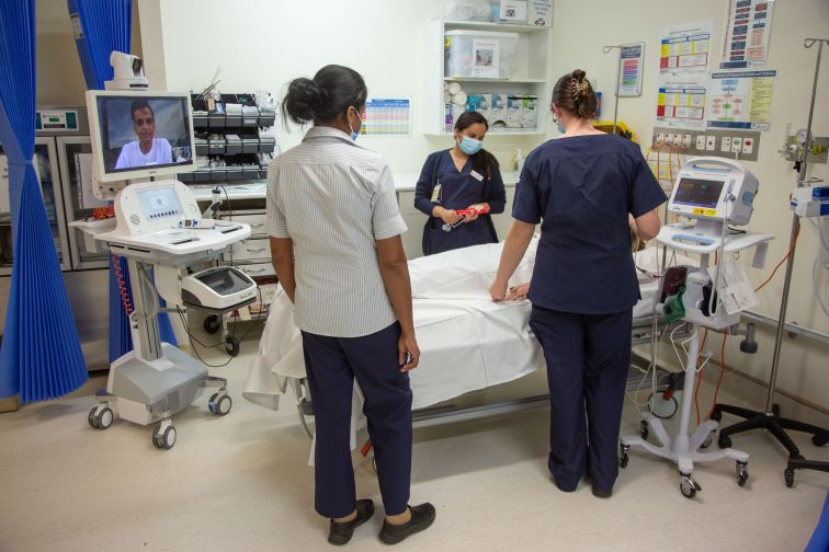 Nurses working with patient bedside under direction of virtual doctor on screen