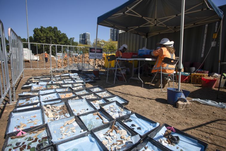 A daytime shot, showing several carefully stacked containers holding findings from an archaeological site, alongside two people wearing high-visibility people working through more containers at a nearby shaded table.