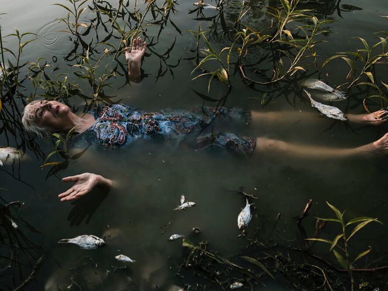 A woman lies in a pond surrounded by dead fish