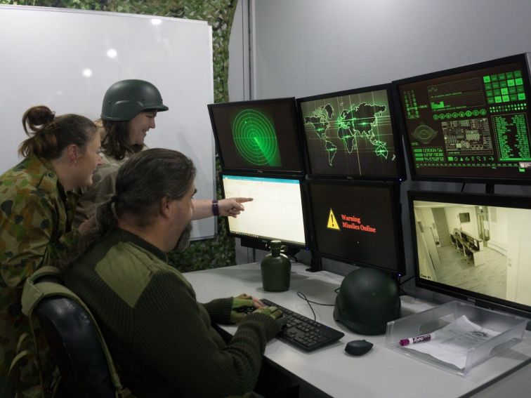 Three people dressed in army gear looking at six monitors