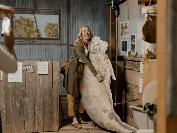 A blonde woman hugs a lifesize cutout of a sheep in a museum.