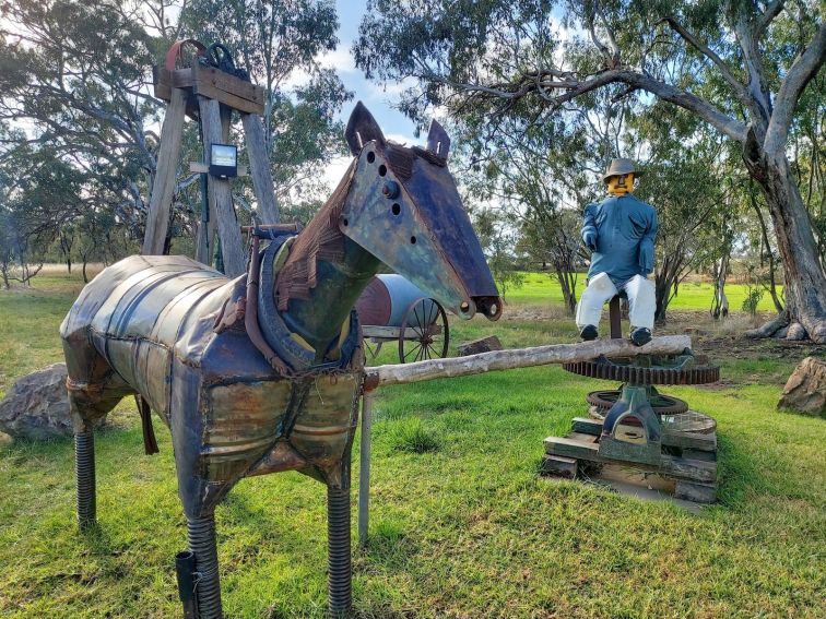 A metal horse sculpture and a man sitting at a water pump in a grassy park.