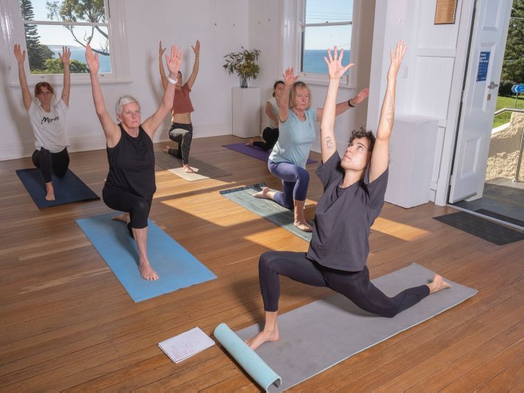 A group of women practice yoga in a sunny hall overlooking the ocean