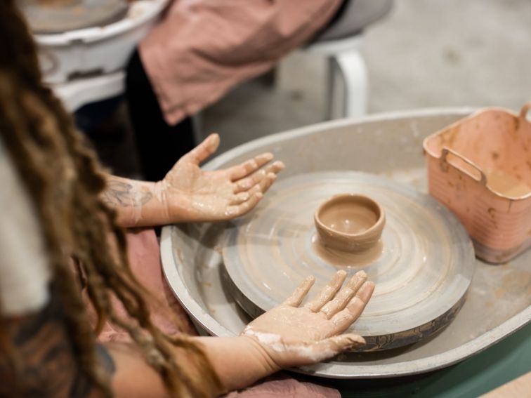 Picture shows a person seated at a pottery wheel with a small clay pot and dirty hands