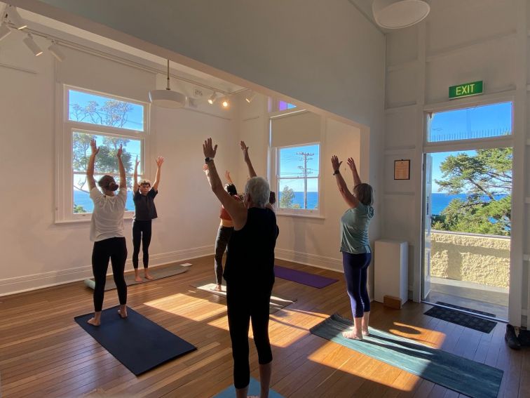 A group of women practicing yoga in a sunny room. The ocean can be seen through the windows.