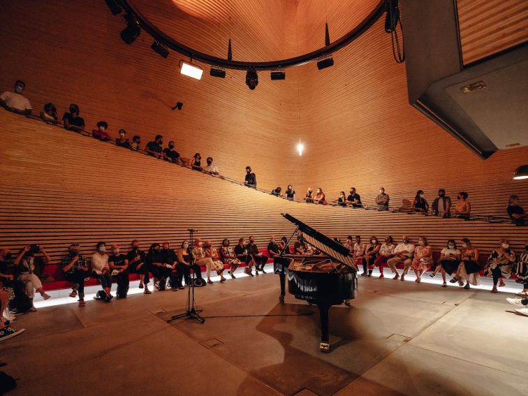 A grand piano sits in a large performance space as people gather