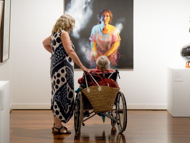 two ladies, one in a wheel chair and one standing admiring a self-portrait by Wendy Sharpe