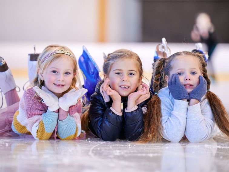 3 young preschool aged girls lying on the ice surface smiling at the camera.