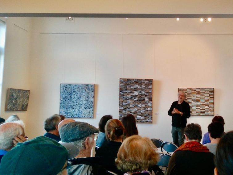An artist speaks to a seated audience with his artworks on the wall behind him.