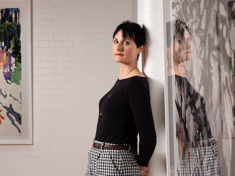 A woman artist leans against a gallery wall and looks at the camera. She has short dark hair.