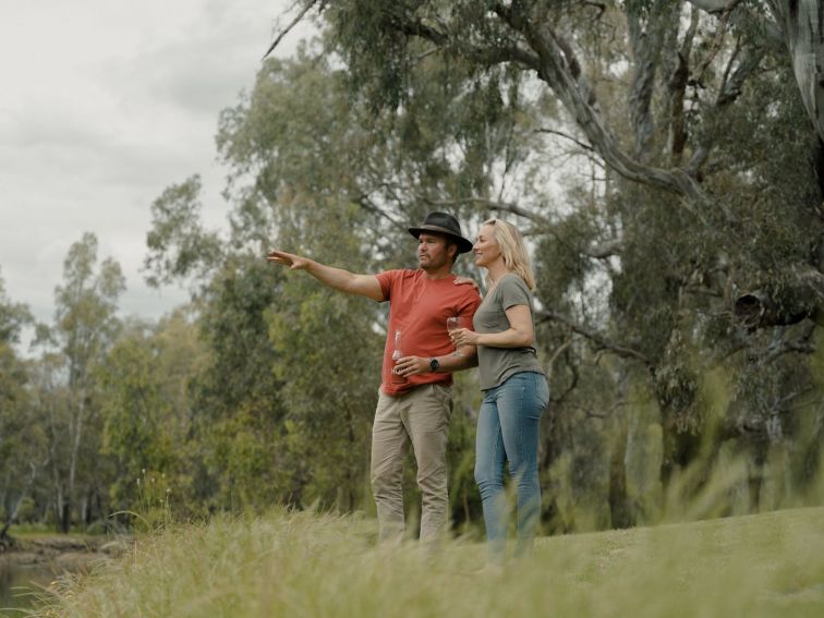 A man and woman stand on the banks of the Murray River, pointing across the river from Howlong.