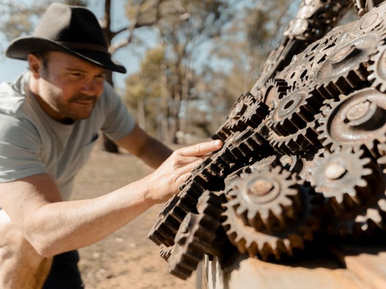 A man rests a hand on iron cogs that form the fleece of a ram sculpture.
